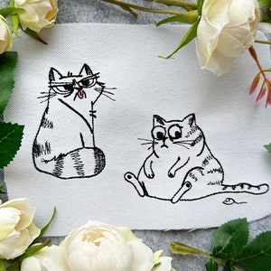 Könnte beinhalten: Ein weißer Stoff mit zwei schwarzen gestickten Katzen. Eine Katze sitzt mit ihrem Schwanz um ihren Körper gewickelt. Die andere Katze liegt mit ihrem Schwanz um ihren Körper gewickelt.