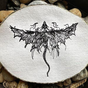 May include: Black embroidered dragon design on a white fabric patch. The dragon is depicted with wings spread and a long tail.