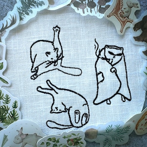 Könnte beinhalten: Ein weißer Stoff mit drei schwarzen gestickten Katzen. Eine Katze ist zusammengerollt, eine andere liegt auf dem Rücken und die dritte steht auf ihren Hinterbeinen.