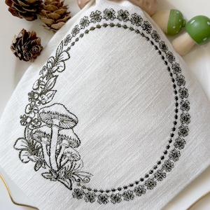 May include: White linen napkin with an embroidered design featuring mushrooms and floral elements. The design is framed by a border of small flowers. Decorative pine cones and mushroom-shaped objects are also visible.