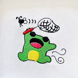 May include: Embroidered design of a green frog with a red hat, holding a net and chasing a black bug. The frog has a yellow belly and is blowing air out of its mouth.