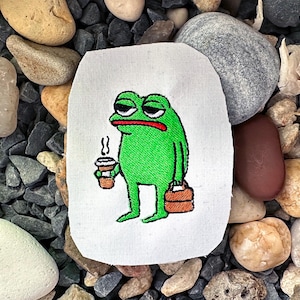 Könnte beinhalten: Grüne gestickte Froschfigur, die eine Tasse Kaffee und eine braune Aktentasche hält. Der Frosch hat einen müden Gesichtsausdruck.