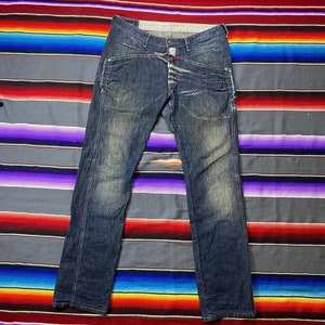 May include: A pair of dark blue denim jeans with multiple layers of fabric sewn together at the waist and front pockets. The jeans have a faded and distressed look.