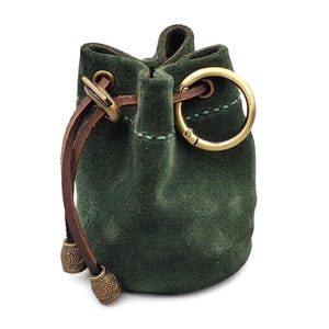 May include: A small, dark green suede drawstring pouch featuring a brown leather cord closure. The pouch is accented with brass-toned metal eyelets, a circular spring-gate ring, and two decorative cylindrical metal beads with engraved star patterns.