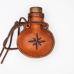May include: A round, tan leather flask featuring a dark brown compass rose design embossed on the front. The flask has a dark brown wooden stopper at the top and a matching leather cord attached through two side loops for carrying or hanging.
