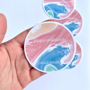 May include: Round sticker with a watercolor design of pink, blue, and green waves. The sticker has the text "Inhale confidence, exhale doubt" written in white.