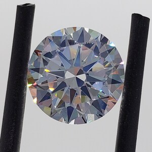 May include: A clear round gemstone with a brilliant cut, held by black tweezers. The gemstone has a rainbow-like effect.