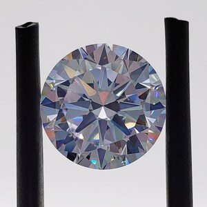 May include: A clear round diamond gemstone with a brilliant cut, held in place by two black metal prongs.