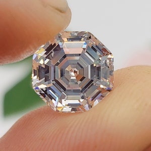 May include: A clear octagon-shaped gemstone with a faceted design. The gemstone is held between two fingers.
