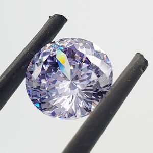 May include: A round, clear, purple gemstone with a faceted cut. The gemstone is held by a pair of black tweezers.