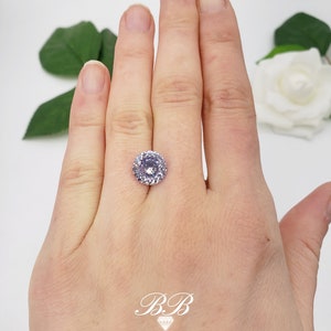 May include: A light purple round gemstone with a halo setting. The gemstone is on a hand.
