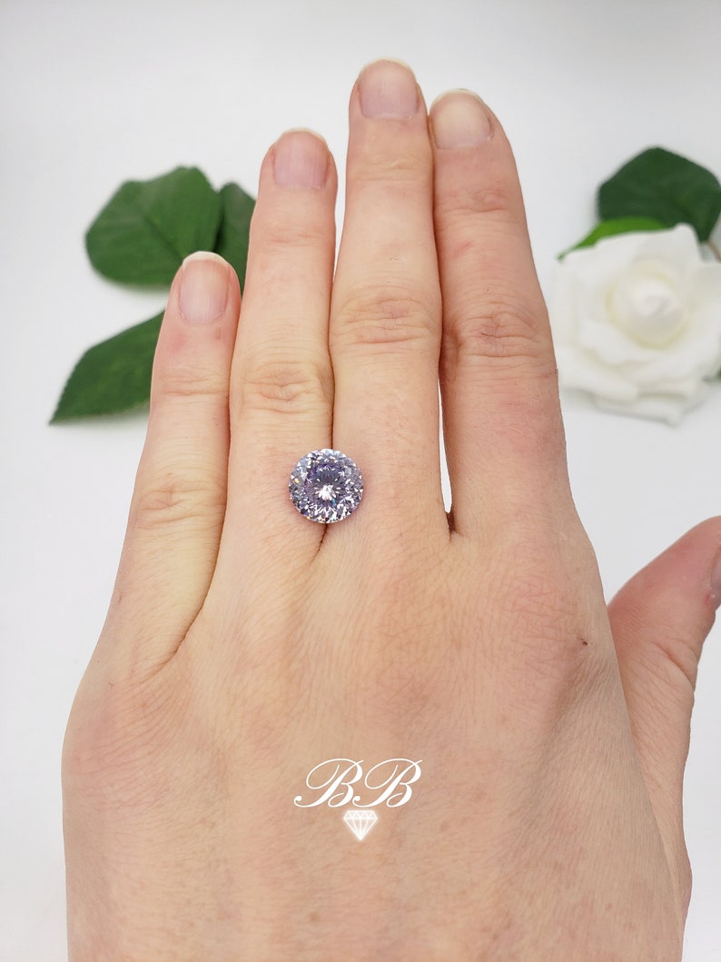 May include: A light purple round gemstone with a halo setting. The gemstone is on a hand.