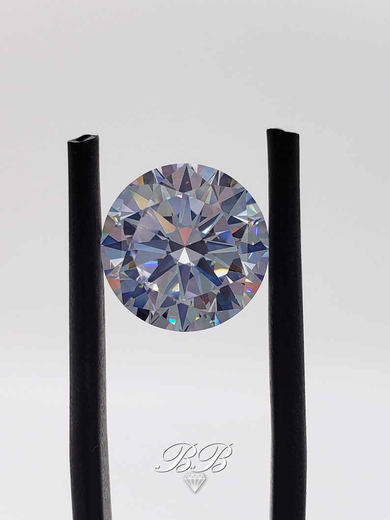 May include: A clear round diamond gemstone with a brilliant cut, held in place by two black metal prongs.
