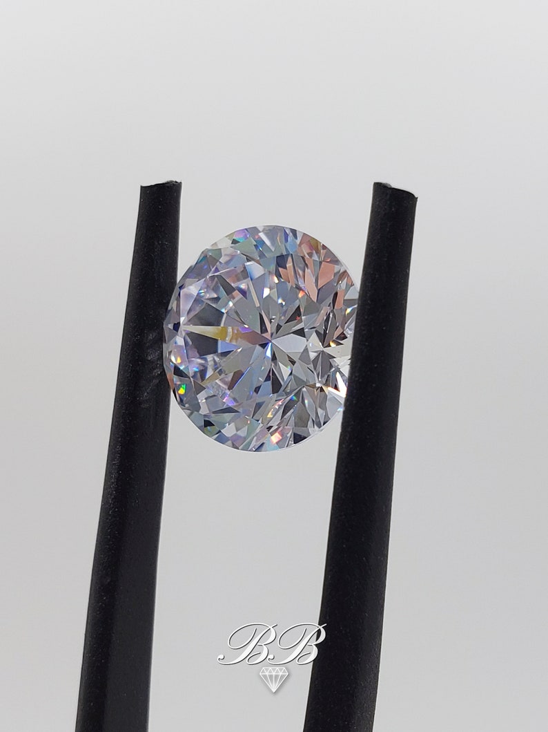 May include: A clear round gemstone with a brilliant cut, held by black tweezers. The gemstone has a rainbow-like effect.