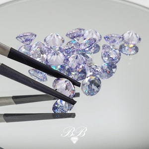 May include: A close-up of a group of iridescent purple gemstones, some of which are being held by a pair of tweezers. The gemstones are round and have a faceted cut.
