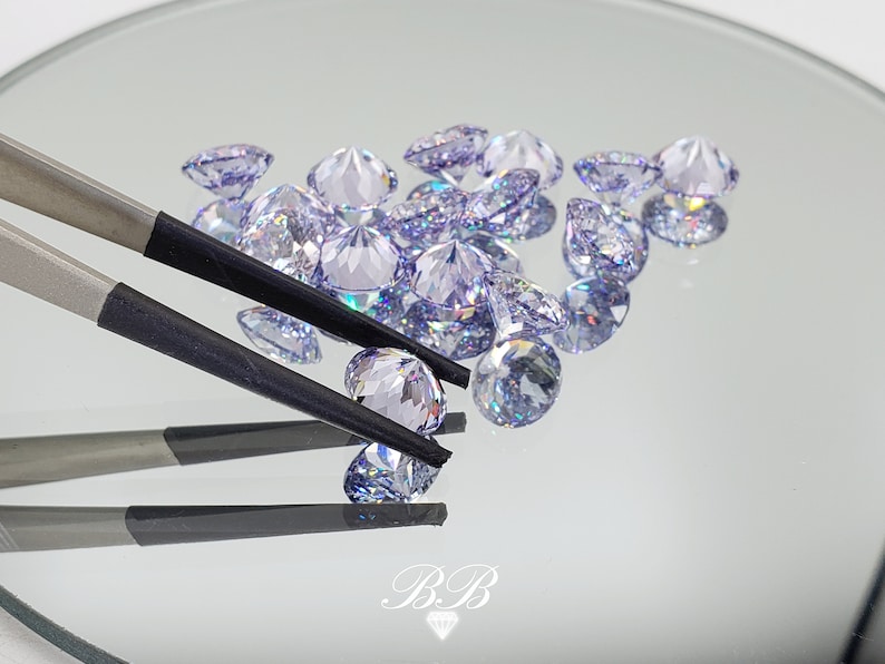 May include: A close-up of a group of iridescent purple gemstones, some of which are being held by a pair of tweezers. The gemstones are round and have a faceted cut.