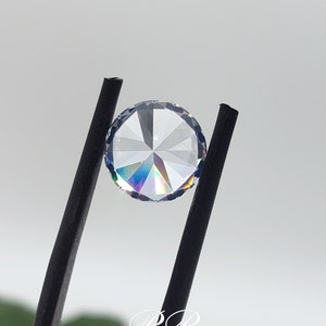 May include: A clear round gemstone with a faceted design, held by black tweezers. The gemstone has a rainbow iridescent effect.