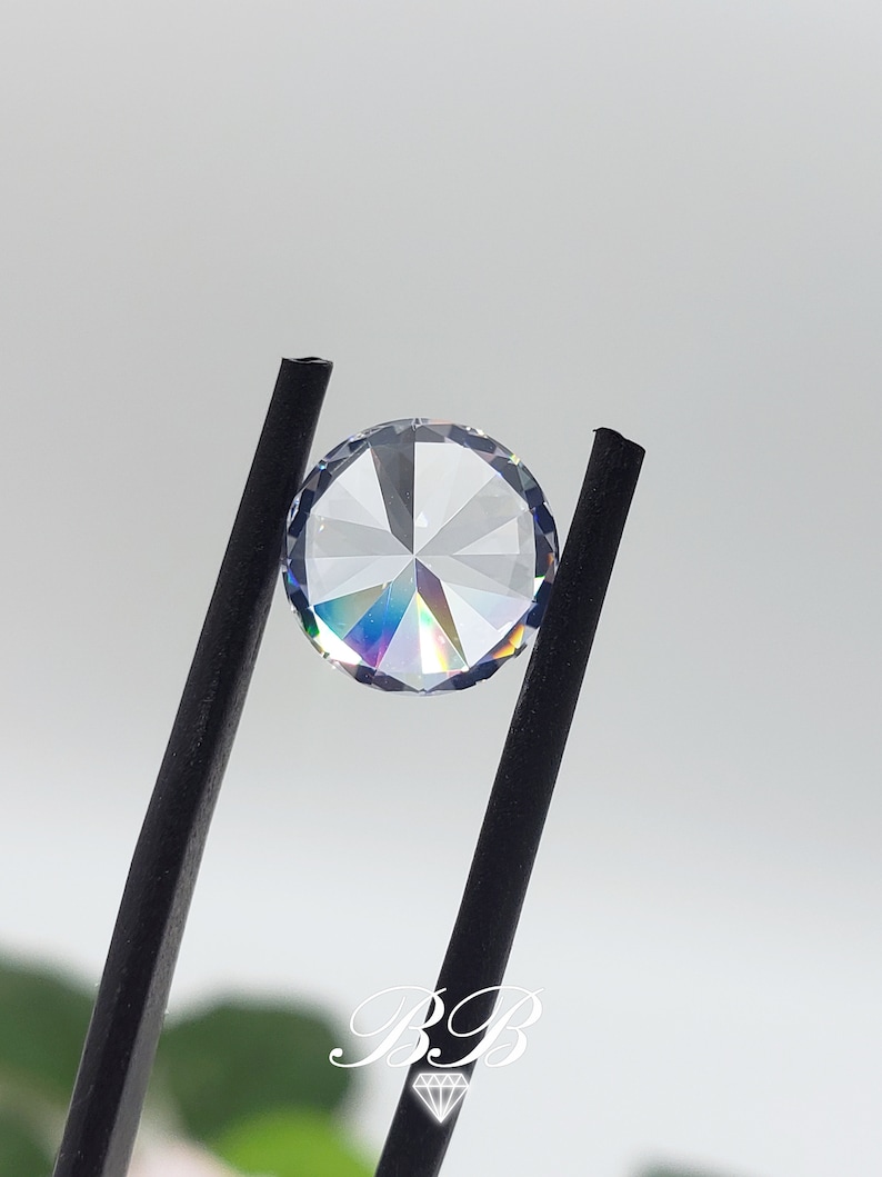 May include: A clear round gemstone with a faceted design, held by black tweezers. The gemstone has a rainbow iridescent effect.