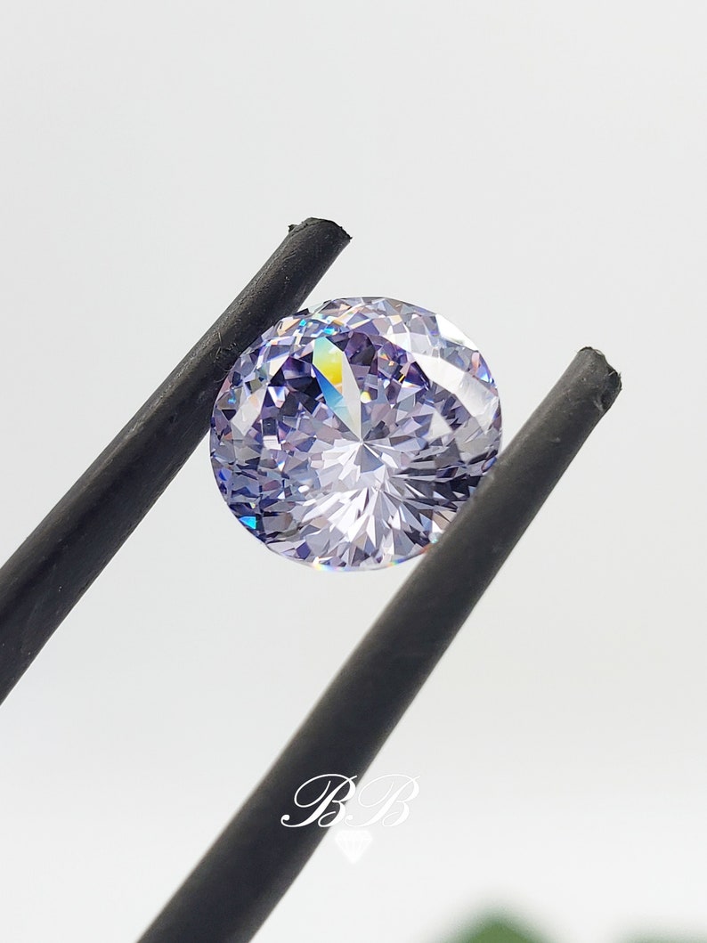 May include: A round, clear, purple gemstone with a faceted cut. The gemstone is held by a pair of black tweezers.