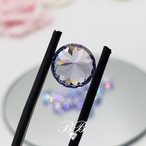 May include: A clear round gemstone with a faceted design, held by black tweezers. The gemstone has a light purple hue and is reflecting rainbow colors.