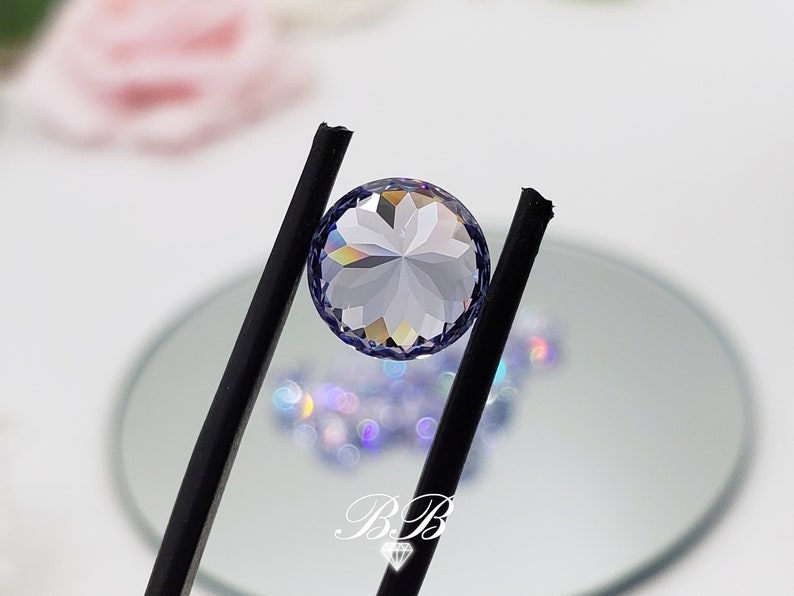 May include: A clear round gemstone with a faceted design, held by black tweezers. The gemstone has a light purple hue and is reflecting rainbow colors.