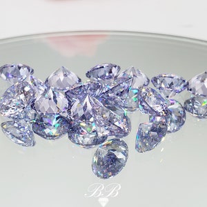 May include: A group of iridescent, light purple, faceted gemstones on a white reflective surface.