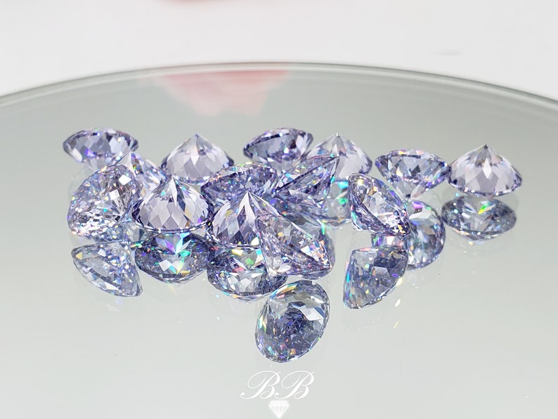 May include: A group of iridescent, light purple, faceted gemstones on a white reflective surface.