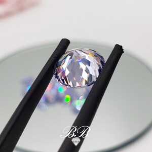 May include: A clear, faceted gemstone with a light purple hue is held by black tweezers. The gemstone is round and has a high degree of brilliance.