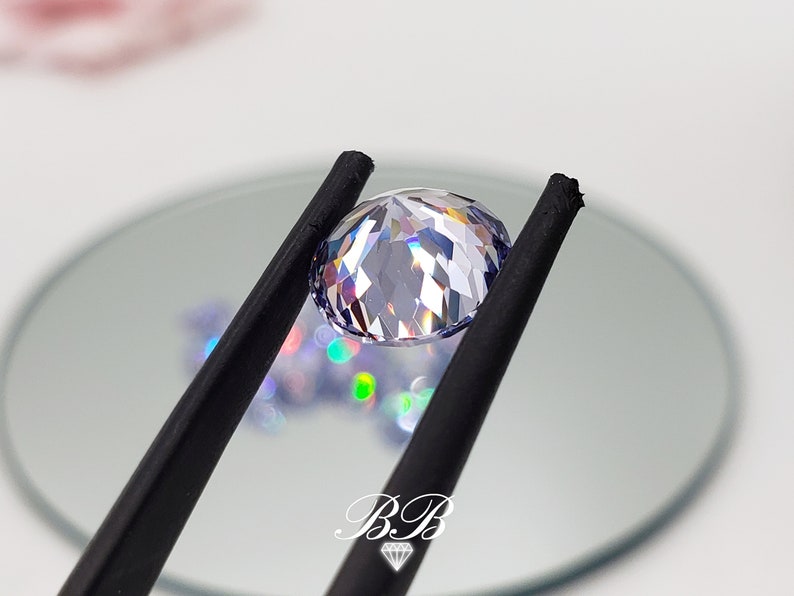 May include: A clear, faceted gemstone with a light purple hue is held by black tweezers. The gemstone is round and has a high degree of brilliance.