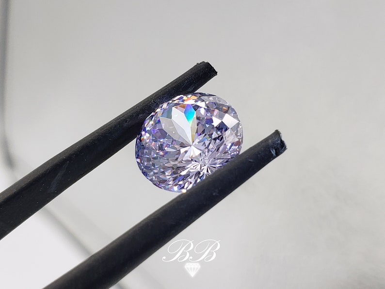 May include: A clear, round, faceted gemstone with a light purple hue. The gemstone is held by a pair of black tweezers.