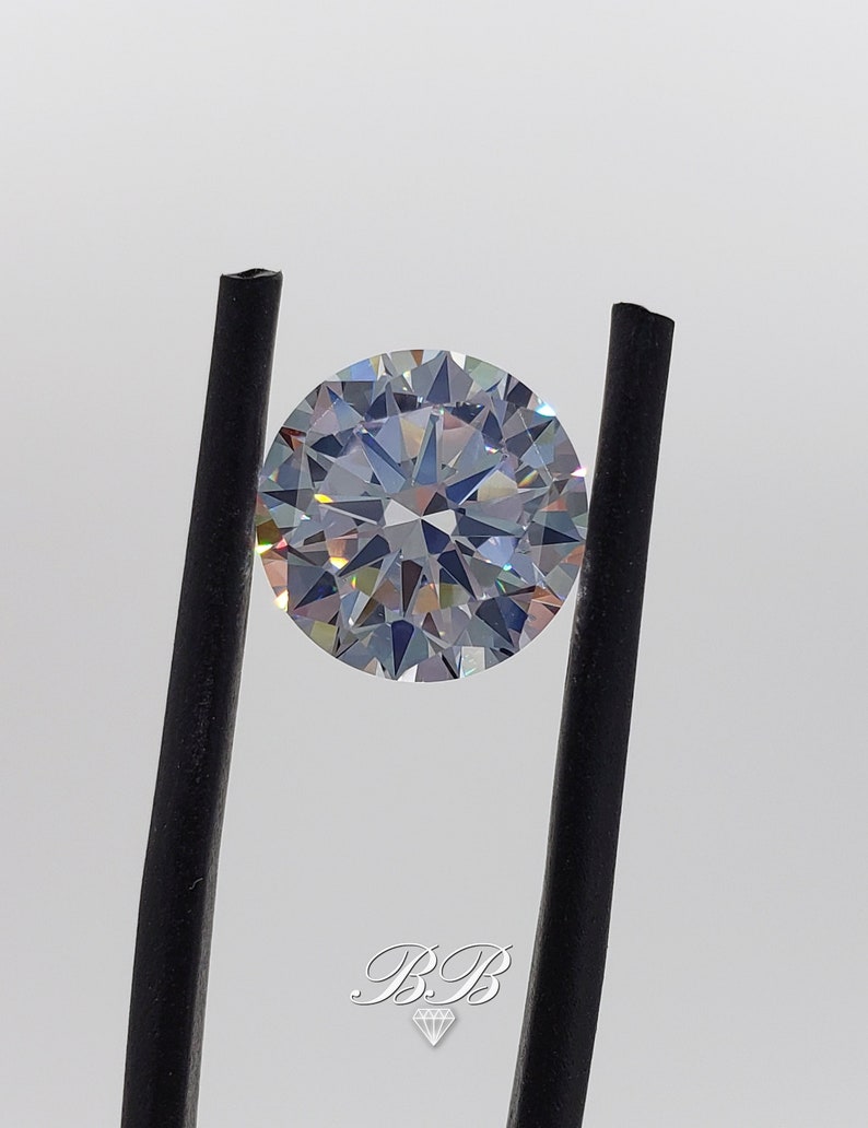 May include: A clear round gemstone with a brilliant cut, held by black tweezers. The gemstone has a rainbow-like effect.