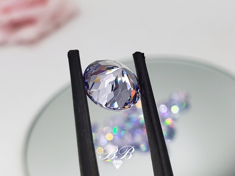 May include: A clear, faceted, oval-shaped gemstone with a light purple hue. The gemstone is held by black tweezers against a white background.