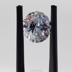 May include: A clear round gemstone with many facets, held in place by two black tweezers. The gemstone is reflecting light, creating a rainbow effect.