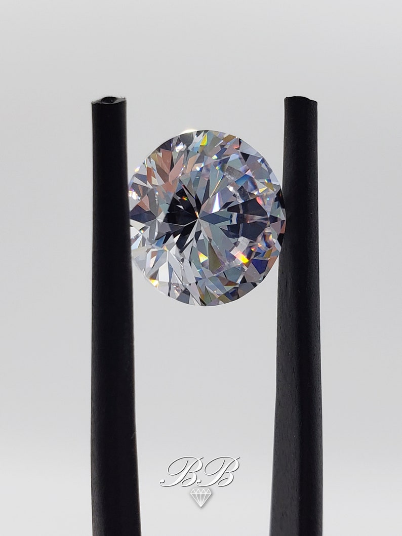 May include: A clear round gemstone with many facets, held in place by two black tweezers. The gemstone is reflecting light, creating a rainbow effect.