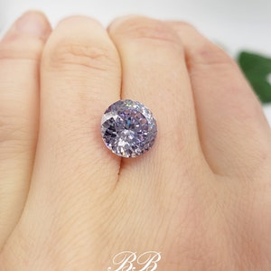 May include: A round, clear, faceted gemstone with a purple hue. The gemstone is set in a ring and is being worn on a finger.