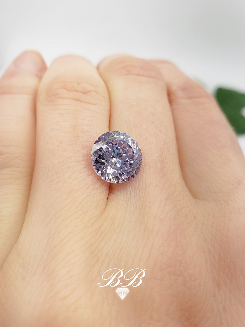 May include: A round, clear, faceted gemstone with a purple hue. The gemstone is set in a ring and is being worn on a finger.