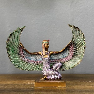 Könnte beinhalten: Eine goldfarbene Figur der ägyptischen Göttin Isis mit ausgebreiteten Flügeln. Die Flügel sind grün, blau und rosa mit goldenen Akzenten. Die Göttin trägt ein violettes und goldenes Kleid mit einer goldenen Krone.