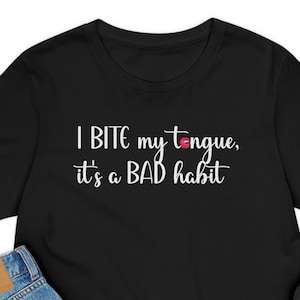 Könnte beinhalten: Schwarzes T-Shirt mit weißem Text, der "I bite my tongue, it's a bad habit" lautet. Das Shirt wird auf weißem Hintergrund mit einem Paar schwarzen Schuhen, einer blauen Jeans und einer beigefarbenen Mütze gezeigt.