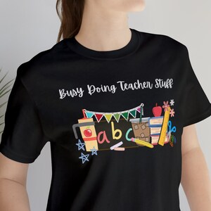 May include: A black t-shirt with the text "Busy Doing Teacher Stuff" and a graphic of school supplies, including an apple, a coffee cup, a chalkboard, and alphabet blocks.
