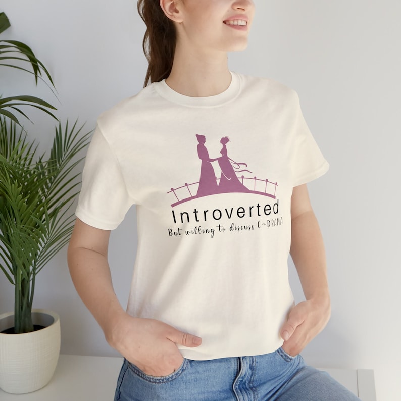 May include: White t-shirt with a pink silhouette of a couple on a bridge and the text "Introverted But willing to discuss C-DRAMA".