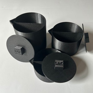 Puede incluir: Tres contenedores de plástico negro con tapas. Cada contenedor tiene un pequeño cuadrado con la palabra "SAC" impresa en él.