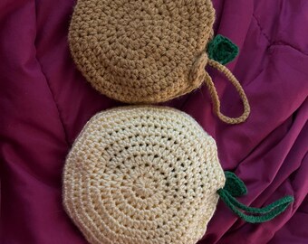 Op de afbeelding: Twee handgemaakte gehaakte zakjes. Eén is lichtbruin, de andere crème. Elk heeft een groen blad en een lus. De ronde zakjes zijn gemaakt van garen. Ideaal voor opslag of decoratie.