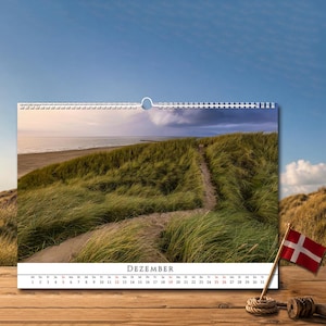 Op de afbeelding: Een wandkalender voor de maand DEZEMBER met een landschapsfoto van een zandpad dat door groen duingras slingert naar een strand en de oceaan onder een bewolkte lucht. Een kleine Deense vlag op een houten stok staat op de voorgrond.