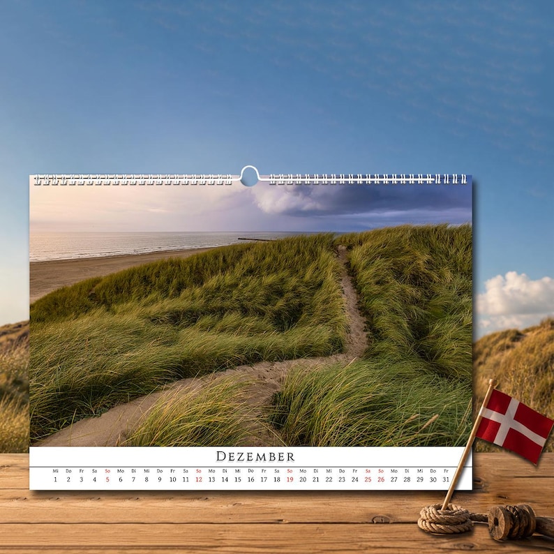 Op de afbeelding: Een wandkalender voor de maand DEZEMBER met een landschapsfoto van een zandpad dat door groen duingras slingert naar een strand en de oceaan onder een bewolkte lucht. Een kleine Deense vlag op een houten stok staat op de voorgrond.