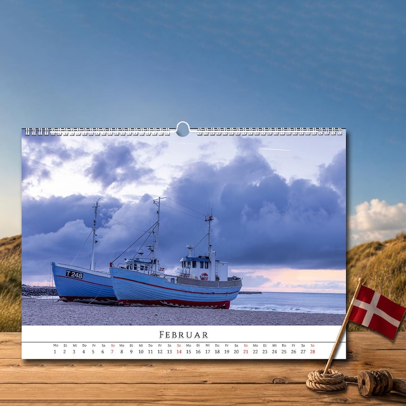 Op de afbeelding: Een wandkalender voor de maand februari met een foto van twee blauwe vissersboten die op een zandstrand liggen onder een bewolkte lucht. Het kalenderraster toont de dagen van de maand onder de afbeelding, met de tekst FEBRUAR bovenaan.