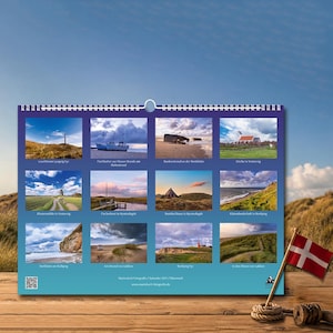 Op de afbeelding: Een wandkalender met twaalf landschapsfoto's van Denemarken, waaronder vuurtorens, kustgezichten en traditionele gebouwen. De kalender heeft een witte spiraalbinding aan de bovenkant en toont de tekst Martin Koch Fotografie Kalender 2023.