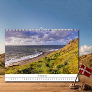 Op de afbeelding: Een wandkalender voor de maand augustus met een landschapsfoto van een met gras begroeide kustklif die uitkijkt over een zandstrand en de oceaan onder een bewolkte lucht. De kalender bevat een kleine Deense vlag aan een houten stok.