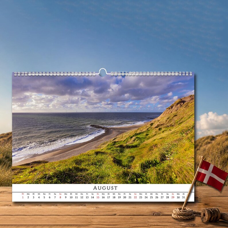 Op de afbeelding: Een wandkalender voor de maand augustus met een landschapsfoto van een met gras begroeide kustklif die uitkijkt over een zandstrand en de oceaan onder een bewolkte lucht. De kalender bevat een kleine Deense vlag aan een houten stok.
