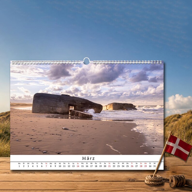 Op de afbeelding: Een wandkalender met een foto van betonnen bunkers op een zandstrand bij zonsondergang met brekende golven. De kalenderpagina toont de maand M&auml;rz met een datumraster onder de afbeelding en een kleine Deense vlag aan een houten stokje.