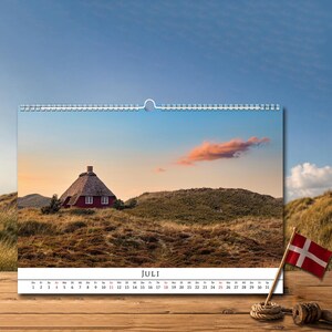 Op de afbeelding: Een wandkalender voor de maand juli met een foto van een rood huis met een rieten dak in een grasachtig duinlandschap onder een zonsondergang. De kalender bevat een datumsraster en een kleine Deense vlag op een houten stokje.